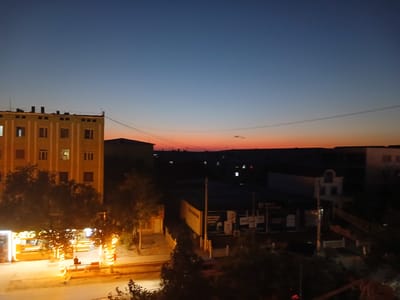 The image captures a dusk scene in Qorowul, Uzbekistan, overlooking a street with buildings and a vibrant sky. In the foreground, a multi-story building with cream-colored walls and numerous windows is visible on the left. The windows on the lower floors are illuminated, casting a warm glow. Trees with lights wrapped around their trunks are positioned in front of this building, creating a festive atmosphere. A street with a paved surface extends across the lower part of the image. A solitary figure is seated on a bench in the illuminated area.
Further back, across the street, there are low-rise commercial buildings, some with illuminated signs and storefronts. One building has text that appears to be in a local script. A streetlamp stands prominently in front of some of these structures. The skyline in the distance is dark, with silhouettes of more buildings and some scattered lights.
The sky is a gradient of colors, transitioning from a deep blue at the top to lighter shades of blue and then a striking orange-pink hue near the horizon, indicative of sunset or twilight. Power lines are visible stretching across the sky. The overall impression is a view of an urban environment at the end of the day, with artificial lighting beginning to dominate as natural light fades.