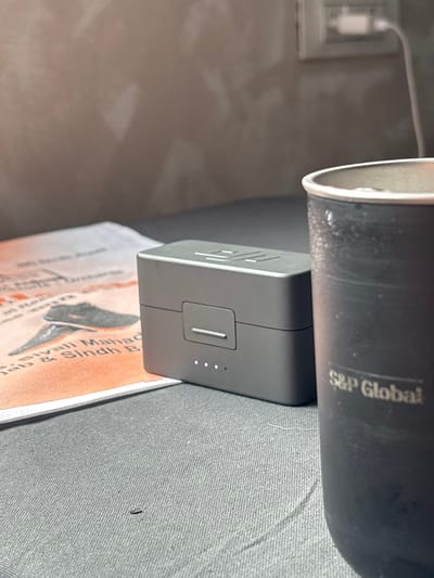 The image shows a close-up of a gray charging case for wireless earbuds, positioned on a dark gray textured surface, likely a table or desk. To the left of the charging case is a white paper with some text and an image of a shoe. To the right of the charging case is a black insulated cup with the logo "S&P Global" visible on its side, containing a beverage with ice or condensation. In the background, a wall with some light fixtures and a white power outlet with a cord is visible. The lighting is soft, creating shadows. The scene appears to be indoors, likely in a home or office setting. There are no people or animals present in the image. The city of Chichoga, India is not depicted or suggested in this image.