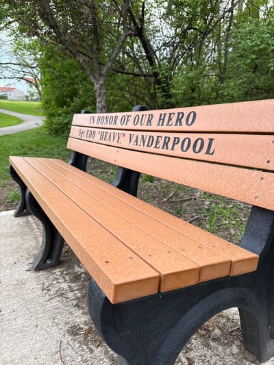 The image shows a park bench in Grove City, Ohio, USA. The bench seat is constructed of tan-colored, wood-like planks with black metal supports. An inscription on the backrest reads "IN HONOR OF OUR HERO Sgt EDD 'HEAVY' VANDERPOOL." The bench sits on a concrete path in a park-like setting with trees and a walking path visible in the background. A building can also be seen in the background.