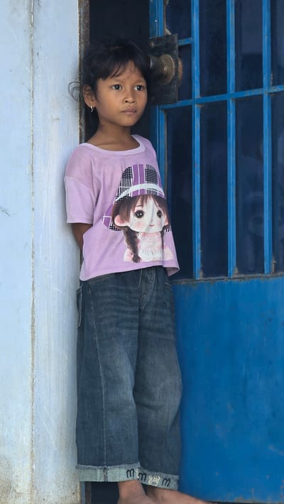 A young girl lies prone on a concrete surface, parallel to a blue metal structure with a window grate. She wears a lilac t-shirt with a large anime girl graphic, jeans with rolled-up cuffs displaying a decorative pattern, and a small earring. Her dark hair is partially covering the concrete. The background includes a section of the blue metal structure with rectangular openings. The scene is located in Ba Ngoi Ward, Vietnam.