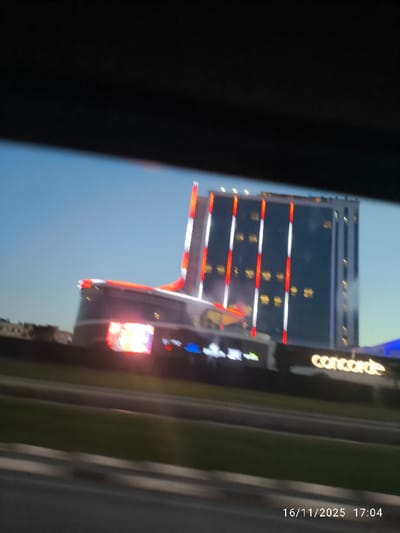 The image displays a blurry, low-angle view of the Concorde Luxury Resort & Casino in Gonyeli, Northern Cyprus, captured from a moving vehicle. The central feature is a modern high-rise building with a dark glass facade, prominently illuminated by vertical red and white light strips. Below the main tower, a curvilinear lower structure is visible, featuring red perimeter lighting and a brightly lit, multi-colored digital display. The name "concorde" is illuminated in white letters on a dark background on the lower portion of the complex. In the foreground, a multi-lane roadway is partially visible, separated by a strip of green landscaping. The sky is clear and light blue, indicating late afternoon or early evening. No individuals or specific vehicles are discernible. The image timestamp is 16/11/2025 17:04.