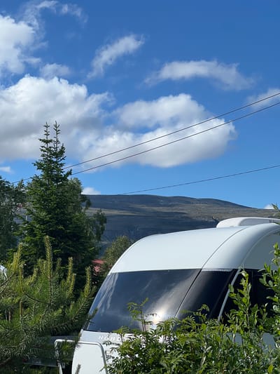 This image shows a close-up view of the side of a white and black recreational vehicle (RV), possibly a caravan or motorhome, with green foliage in the foreground. The RV appears to be parked outdoors, with a large, dark, and mountainous landscape visible in the background, suggesting a natural setting. The sky above is blue with scattered white clouds, indicating fair weather, likely during the daytime. Several dark, thin cables stretch across the sky in the middle ground.

The RV has a sleek, modern design, with a curved white section and a contrasting dark, possibly tinted, panel. Some greenery is visible peeking out from behind the RV, and more prominently in the foreground, with branches and leaves obscuring parts of the vehicle. The sunlight reflects off the shiny surface of the RV, creating a bright glint.

Although the image is a still photograph and no activity is explicitly depicted, the presence of the RV and the natural landscape implies a travel or recreational setting. The image is oriented vertically, as if taken with a phone held in portrait mode, and then rotated. There is some text visible on the side of the RV, partially obscured by foliage and the curve of the vehicle. The visible text appears to be "HOBE."

The overall impression is one of a scenic outdoor location, possibly a campsite or a viewpoint, where the RV is situated amidst nature. The bright sunlight and blue sky suggest a pleasant day, ideal for outdoor activities. The location context of Lakselv, Norway, further supports the idea of a natural, possibly mountainous or fjord-like, environment.
