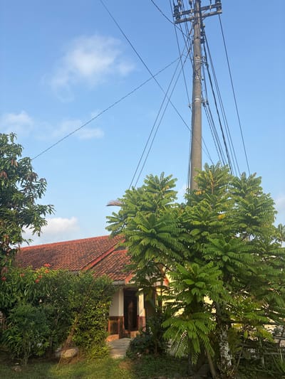 A power pole with numerous cables stands tall in front of a house with a red tile roof, surrounded by lush green trees and vegetation under a bright blue sky with scattered clouds. The house appears to be a single-story dwelling with a visible entrance and decorative stone accents. The scene captures a residential area in Majenang, Indonesia, likely during daylight hours given the bright illumination.