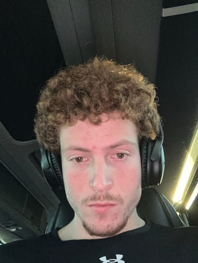A fair-skinned person with curly auburn hair is wearing black headphones around their neck. The individual has light brown eyes, a stubbled beard, and a few blemishes on their face. They are wearing a black shirt with a visible logo on the right side. The background appears to be the interior of a vehicle, with dark panels and a light reflecting overhead. The image is rotated.