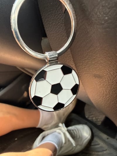 A close-up shot captures a shiny silver keychain featuring a circular charm with a classic black and white soccer ball design. The keychain's large ring is prominent on the left, while the soccer ball charm, depicting traditional hexagonal and pentagonal panels, is centrally placed and gleams under the light.
The scene appears to be set inside a car. A person's light-skinned leg and foot are visible in the lower right portion of the frame. The foot is clad in a white low-cut sock and a white lace-up sneaker, suggesting the person could be a passenger or a driver resting their foot.
The background consists of dark, textured surfaces, typical of a car interior, possibly parts of a seat, dashboard, or floor mat. Hints of car pedals can be discerned in the deeper background towards the bottom right, further confirming the car setting. The lighting indicates it is daytime. No visible text is present in the image. The location context for this image is Radinovo, Bulgaria.