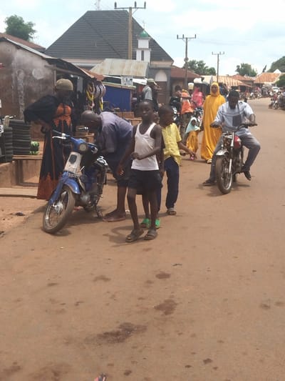 The scene is a street in Lokoja, Nigeria (7.404, 7.632). There is a woman wearing a black and orange robe standing next to a blue motorcycle. A man is bent over, working on the motorcycle. Two boys are standing near the motorcycle, and two more are walking away from the camera in the distance. There is a man riding a black motorcycle in the distance. In the background there is a mosque and buildings along the road. The street is paved and there is a lot of dust. The sky is clear with few clouds. There are many other people, bikes and cars walking in the background.  The scene is a typical street scene in Lokoja, Nigeria.