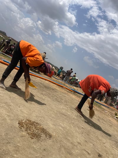 Two individuals are sweeping a dirt surface with brooms, situated in Maiyanga, Nigeria. The sweepers, both barefoot, are wearing orange tops and dark pants. The person closer to the camera is also wearing a striped hat and holding a yellow object. An orange and blue hose lies on the ground. A scattering of small, brown objects is visible on the dirt surface. In the background, a group of people and some vehicles are present. The sky is partly cloudy.