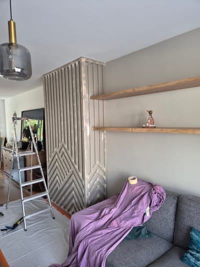 This is a medium shot photograph, taken from a low angle, of a living room undergoing renovation. The room's dominant feature is a slatted wooden wall panel with a geometric pattern, painted a light grey. Above this, two parallel wooden beams run horizontally across the frame, attached to a light grey wall which extends to the ceiling.
In the upper right quadrant of the image, a portion of a dark grey textured sofa is visible, draped with a large piece of light purple fabric. A roll of masking tape is attached to the fabric. To the left of the sofa, a small, decorative item resembling a vintage toy car is placed on one of the wooden beams.
In the lower part of the frame, a silver ladder is positioned in front of the slatted wall, with a white sheet spread on the floor beneath it, suggesting painting or decorating activities. A television screen is visible behind the ladder, displaying an outdoor scene with greenery. A modern pendant light with a smoked glass shade and brass accents hangs from the ceiling on the left side of the image.
The overall impression is that of a home improvement project in progress. The lighting suggests daytime, with natural light likely coming from a window out of frame. There is no visible text in the image. The setting appears to be a contemporary residential interior in Schengen, Luxembourg.