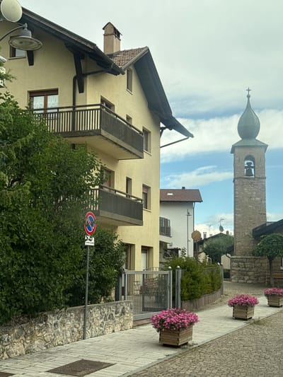 In Fai della Paganella, Italy, a street scene unfolds with a pale yellow building dominating the left side of the frame, featuring two dark-railed balconies. Adjacent to the building is a dense green hedge. A street sign, depicting a red circle with a diagonal bar and a blue symbol, is mounted on a pole beside the hedge. The street pavement is made of light-colored rectangular stones. Beyond the building, a stone church tower with a green dome and a bell is visible. The background shows a cloudy sky. In the foreground, three wooden planters filled with vibrant pink flowers are placed along the street.