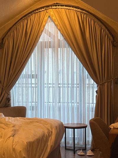 The image shows the interior of a room, possibly in a hotel or private residence. The focal point is a large window covered with sheer white horizontal blinds, partially obscured by ornate, heavy tan-colored drapes. The drapes are gathered at the top and sides, creating a diamond-shaped opening that reveals the blinds.
To the right of the window is a small round table with a dark metal frame and a light-colored wooden top. Next to the table are two pairs of white slippers. A portion of an upholstered chair is visible, partly covered with a rumpled white blanket or bedspread. The lighting in the room appears soft and diffused, creating a warm ambiance. The interior could be located in Ürgüp, Turkey, but there is nothing in the image that confirms it, it would be speculation.