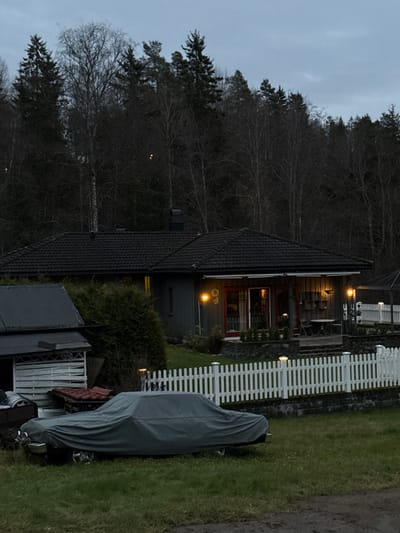 The image depicts a residential property at dusk or dawn in Jessheim, Norway. A dark gray, single-story house with a dark tiled roof is the central subject, featuring vertical siding and a visible chimney. Its front area includes a patio or deck with a small table and two chairs, illuminated by two heart-shaped wall-mounted lights and interior lighting visible through red-framed glass doors and windows. A white picket fence, topped with small white lights, runs along the property's edge, situated on a low, dark stone retaining wall.

To the left of the house, a smaller, dark-roofed outbuilding or shed with light-colored slatted siding and stored items underneath is present. In the immediate foreground, a vehicle is parked on grass, covered entirely by a gray tarp, with portions of its wheels visible. The surrounding environment consists of green grass and a dense background of trees, including both tall evergreens and bare deciduous trees, suggesting a temperate climate and possibly late autumn or winter. The sky above is overcast and pale. No individuals are visible in the scene.