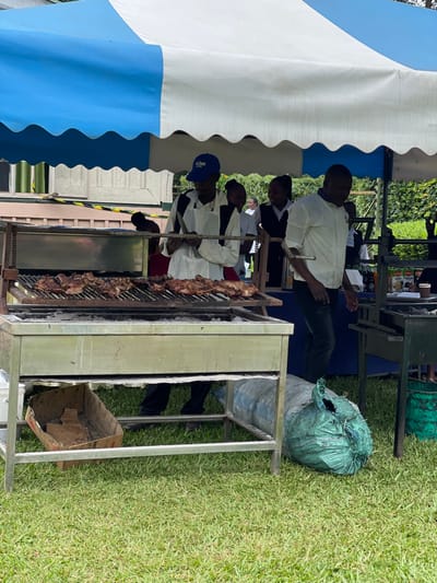The image, rotated ninety degrees clockwise, captures an outdoor catering event or gathering taking place on a lush green lawn in Makuyu, Kenya. A large white and blue striped tent provides shade on the left side of the frame, under which several men, appearing to be catering staff or chefs, are at work.
The central activity revolves around a long, heavy-duty metal grill or rotisserie, which is laden with numerous cuts of meat, likely being prepared as 'nyama choma,' a popular Kenyan barbecue style. Hot coals are visible beneath the grill, indicating active cooking. At least four men are directly involved with the grilling process. One man in a white collared shirt and dark trousers stands to the right of the grill, seemingly overseeing the operation. Another man, wearing a blue baseball cap with faint, unreadable text and a white uniform, leans over the grill, tending to the meat. Other uniformed individuals are visible further back under the tent, with a display table showing partially obscured food items or products in the background.
Near the grilling station, a large, dark green sack, possibly containing charcoal or waste, rests on the grass. A metal table with cardboard boxes underneath is also present. The scene is illuminated by bright natural daylight, suggesting a daytime event. The atmosphere appears focused and industrious, centered on the preparation of a large quantity of barbecued meat for an audience or guests. No specific emotions are clearly discernible from the participants, who are concentrated on their tasks.