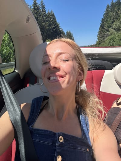 A blonde woman with her eyes closed and a wide smile is photographed from the chest up, taking a selfie in the passenger seat of a convertible. She is wearing a denim jumpsuit and large hoop earrings, and a seatbelt is visible across her chest. The top of the convertible is down, and a sunny, tree-lined landscape is visible in the background. The scene appears to be a road in the Furtwangen im Schwarzwald, Germany region.