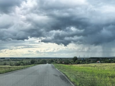 The media file shows a paved road leading through a rural landscape under a dramatic, cloudy sky. The scene is a wide shot, capturing an open vista with fields of green grass on either side of the road. In the distance, there are scattered trees, some small buildings with red roofs, and power lines. The sky is filled with dark, heavy storm clouds, with a visible rain shower falling in the far right of the frame. The overall mood is somber and perhaps a bit foreboding due to the impending or ongoing rain. The lighting suggests it is daytime, but the overcast conditions create a muted, diffused light. The location cues, such as the power lines and the style of the buildings, are consistent with a rural setting in Russia. There is no text visible in the image. The focus is on the natural landscape and the approaching weather.