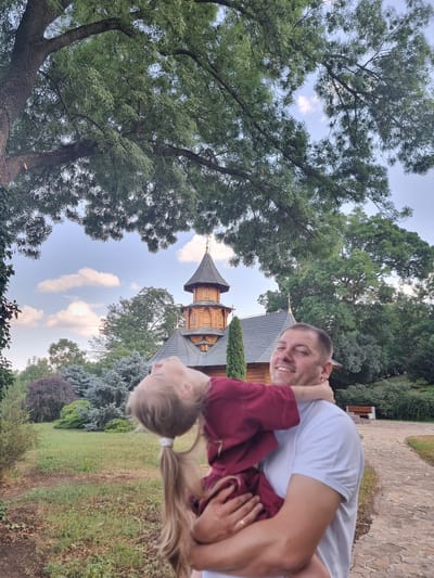 A man and a young girl are depicted in a playful embrace, with the girl being held upside down. The man, with short hair and a white t-shirt, smiles broadly at the camera, his head tilted back. The girl, with long blonde hair tied in a ponytail, wears a maroon top and looks upwards. They are positioned in front of a distinctive wooden building with a conical roof, reminiscent of a church or historical structure. The surrounding environment is lush and green, featuring mature trees with dense foliage and a grassy area. The sky above is blue with scattered white clouds, suggesting it is daytime, likely late afternoon or early evening given the soft lighting. The stone pathway behind the man and to his right hints at a park or a well-maintained outdoor area. The overall atmosphere is one of joy and lightheartedness, captured in a natural, outdoor setting. The location is identified as Hagimus, Moldova. There is no visible text in the image.
