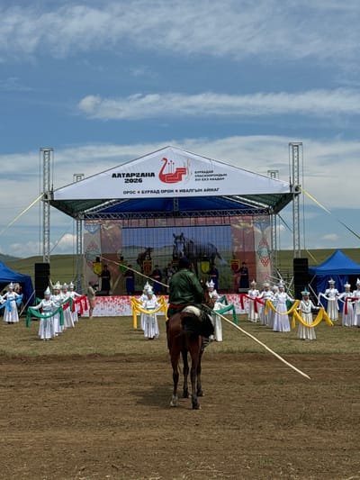 This image depicts an outdoor cultural event in Orongoi, Russia, under a partially cloudy blue sky. In the foreground, a person on horseback faces away from the camera, holding a long wooden pole. Behind them, a group of individuals in white traditional clothing with colorful sashes perform on a grassy field. They are arranged in two rows, with women in the front holding green and red sashes, and women in the back holding yellow and white sashes. A stage with a large white banner is set up behind the performers, featuring Cyrillic text and graphics. The banner displays the text "АЛТАРГАНА 2026" and "БУРЯАДАЙ УЛАСХООРОНДЫН XVI ЕХЭ НААДАН". In the background, rolling hills and some blue tents are visible. The ground is covered in dirt and grass, suggesting a rural setting. The event appears to be a celebratory gathering or festival.