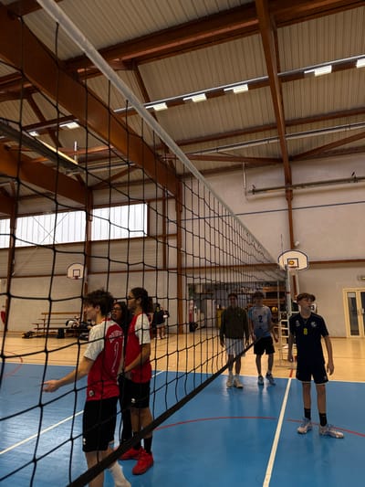 The image depicts an indoor volleyball court within a sports gymnasium in Sennecey-le-Grand, France. A black volleyball net spans horizontally across the middle ground, separating two groups of individuals on a blue synthetic court marked with white lines. The gymnasium features a high, peaked corrugated metal roof supported by dark wooden beams and steel trusses. Numerous linear fluorescent lights illuminate the space from above, complemented by natural light from high rectangular windows along the side walls.
On the left side of the net, a young male in a red and white jersey with "1" visible stands in the foreground, looking left with a slight smile, his left arm extended. Behind him, a female with glasses and dark hair is flanked by another male with long dark hair, also in a red and white jersey. Several other individuals are partially visible further back on this side of the court, near bleachers and a basketball hoop.
On the right side of the net, three young males are positioned on the court. From left to right: one wears a dark green long-sleeved top and white shorts; another wears a light blue and white jersey with "2" visible and black shorts; and the third, in a dark blue jersey with "8" visible and dark blue shorts, looks forward. A basketball hoop is visible in the background on this side of the court, along with shelving containing sports equipment and a door. The individuals are dressed in athletic wear and appear to be engaged in or observing a volleyball session.
