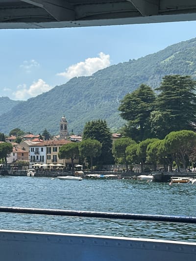 The image depicts a scenic view of the waterfront in Tremezzina, Italy, under a clear blue sky with scattered clouds. In the foreground, a dark railing and a blue metallic structure partially obscure the view of a calm body of water, likely Lake Como, which is a prominent feature of Tremezzina.
On the opposite shore, a picturesque Italian village is visible. Buildings with terracotta roofs and light-colored facades line the waterfront, interspersed with lush green trees. A tall church tower with a clock face stands out amongst the architecture, characteristic of many Italian towns. In front of the buildings, outdoor seating areas with umbrellas suggest cafes or restaurants. Several boats are moored along the shore and floating on the water, indicating a bustling aquatic environment.
Behind the village, a majestic, densely forested mountain rises, its slopes covered in vibrant green foliage, suggesting a natural backdrop. The overall impression is one of a tranquil and idyllic lakeside setting, capturing the essence of Tremezzina's charm.