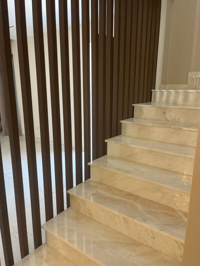 The image shows an interior view of a modern staircase with marble steps and a slatted wooden partition. The partition consists of vertical, dark brown wooden slats arranged closely together. Behind the partition, a white wall and what appears to be a window or opening with a light-colored curtain are visible. The staircase itself is made of polished marble with a creamy beige color and distinct veining patterns. The steps are wide and well-lit, reflecting the ambient light. The overall impression is one of a luxurious and contemporary interior design. The scene appears to be in a residential or commercial building located in Moni Community, Cyprus. There are no people or animals visible in the image.