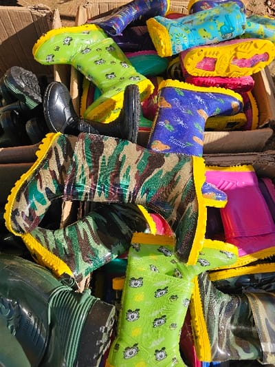 A chaotic pile of colorful rubber boots fills the frame. Patterns vary from camouflage and solid colors to bright blues, greens, and pinks adorned with cartoon characters. Yellow soles and accents are common, contrasting against the various uppers. The boots are piled haphazardly within a cardboard box, creating a jumbled and visually stimulating arrangement.