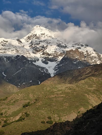 The image depicts a majestic, snow-capped mountain peak dominating the frame. The mountain is partially shrouded in dramatic clouds, with patches of blue sky visible above and through the cloud cover. Sunlight illuminates the snowy slopes, highlighting their rugged texture and the dark, rocky outcrops.
In the foreground, rolling hills covered in sparse green vegetation and scattered rocks recede into the middle ground. The terrain in the foreground appears to be a lower elevation, contrasting with the snow-covered heights. The sunlight casts shadows across these lower slopes, suggesting a time of day when the sun is at an angle, possibly morning or late afternoon.
The scene evokes a sense of awe and the grandeur of nature. The overall atmosphere is one of serene, wild beauty. There are no visible people, objects, or text in the image. The location is identified as Stingri, India, suggesting a mountainous region within that area.