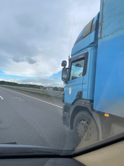 The image shows a blue Scania truck traveling on a highway. The photo was taken from inside another vehicle, likely a car, moving alongside the truck. The highway has a yellow line marking the lanes. A roadside guardrail and some trees are visible beyond the truck. Buildings are visible in the distance. The sky is cloudy. The truck has a Scania logo and the word "Scania" visible on the cabin. Based on the location, Бавыкино, Russia, this scene is likely taking place on a highway in that area.