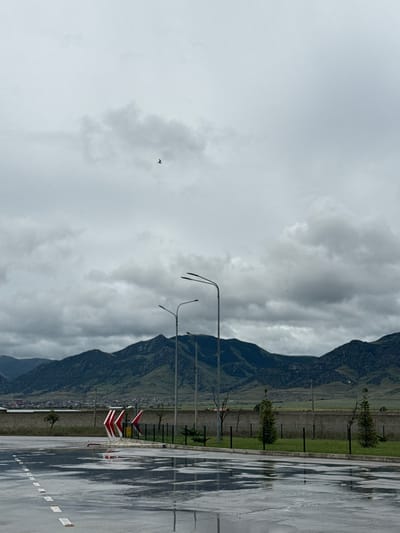 This outdoor scene, taken in Korkmaskala, Russia, captures a vast, overcast landscape with evidence of recent rain. The image is composed of a foreground featuring a wet paved surface, a mid-ground with various structures and greenery, and a background dominated by mountains under a cloudy sky.

In the foreground, a wide, wet asphalt or concrete area, possibly a parking lot or road, is visible, reflecting the grey sky and creating shimmering patterns. White dashed lines are painted on this surface.

The mid-ground includes several tall streetlights or lampposts with multi-arm fixtures. To the right, prominent red and white chevron signs indicate a curve or hazard. A strip of green grass and a low brick wall run parallel to the paved area, behind which a few small trees or bushes are scattered. Faint outlines of buildings or a small settlement can be seen at the base of the mountains in the distance.

The background is dominated by large, imposing mountains, covered in green vegetation, stretching across the entire frame. The sky above is completely overcast with thick, grey clouds, suggesting a dull or rainy day. A single, small dark bird is captured in flight against the expansive grey sky. No people are visible, and there is no discernible text. The overall mood is quiet and somewhat somber, influenced by the cloudy weather and natural landscape.