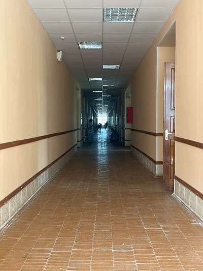 The image shows a long, narrow corridor with light beige walls and a brown tiled floor. Fluorescent lights are installed in the suspended ceiling. Along the walls, at mid-height, there is a dark brown wooden trim. On the left wall, closer to the foreground, a white smoke detector and a white speaker are visible. Further down the corridor, on the right side, a wooden door with a silver handle is present. A red fire extinguisher box is mounted on the wall in the distance on the right. Several individuals are present in the corridor, some standing and others appearing to be sitting or crouched. The overall impression is of an interior hallway, possibly in a public building or institution. The location of Караваево, Russia is not explicitly visible in the image.