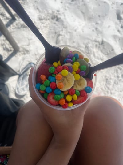 The image captures a close-up, top-down view of a person holding a clear plastic cup filled with a colorful snack, set against a sandy background.
In the foreground, a person's hands and arms are visible, holding the transparent plastic cup. The skin tone is light to medium. The person's legs, possibly clad in shorts or swimwear, are visible in the upper right and lower right portions of the frame.
The cup is overflowing with various toppings. Prominently featured are numerous circular slices of banana, which are light yellow in color. Alongside the bananas are bright red slices of strawberry. Interspersed throughout the fruit, and covering a significant portion of the surface, are small, candy-coated chocolates (resembling M&M's) in a vibrant array of colors including red, orange, yellow, green, blue, and purple. The base of the snack in the cup appears to be a darker, reddish-purple mixture, possibly açai or another berry blend, though it is mostly obscured by the toppings. Two dark-colored plastic spoons are inserted into the cup, one pointing towards the upper left corner and the other towards the lower left.
The background consists primarily of light-colored, textured sand, strongly suggesting a beach environment. The sand shows subtle variations in light and shadow, indicating its granular nature and some undulations. In the lower left, a blurry, dark, linear object is faintly visible, possibly part of beach furniture like a chair leg.
The lighting is bright and natural, indicating it is daytime, likely outdoors under sunny or overcast skies that still provide ample illumination. The overall scene conveys a relaxed and leisurely atmosphere, likely depicting someone enjoying a treat at the beach. There is no visible text in the image.