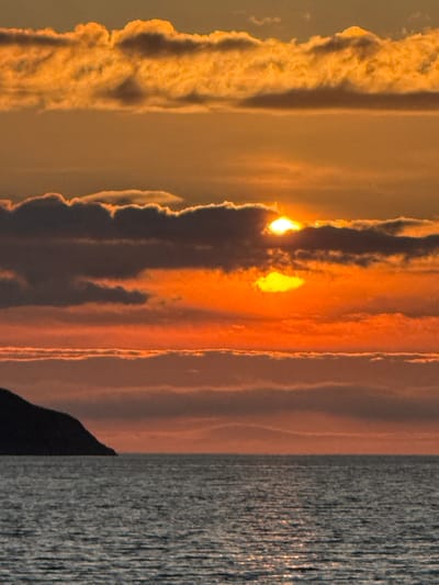 A dramatic sunset unfolds over the Barents Sea in Teriberka, Russia, casting the sky in hues of orange, yellow, and deep red. Clouds, illuminated from behind, create a textured and layered effect across the horizon. Partially obscured by these clouds, the sun appears as two bright, glowing orbs. The water below reflects the vibrant colors of the sky, with shimmering light dancing on its surface. To the left, the dark silhouette of a rocky shore or small island juts out into the sea, its form softened by the low light. The overall scene is one of natural beauty and the raw power of an Arctic sunset.