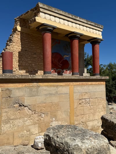 The image shows a section of the Palace of Knossos, an archaeological site located in the city of Heraklion, Greece. In the foreground, large weathered stones are scattered on the ground. The midground features a reconstructed section of the palace, characterized by a bright yellow entablature with decorative circular motifs, supported by three prominent red columns with black bases and capitals. Behind the columns, a portion of a fresco depicting a red bull can be seen. The walls are constructed of rough-hewn, tan-colored stone blocks, with areas of visible damage and missing sections. The background reveals more stone structures of the palace and lush green trees under a clear blue sky. The scene is illuminated by bright sunlight, casting distinct shadows.
