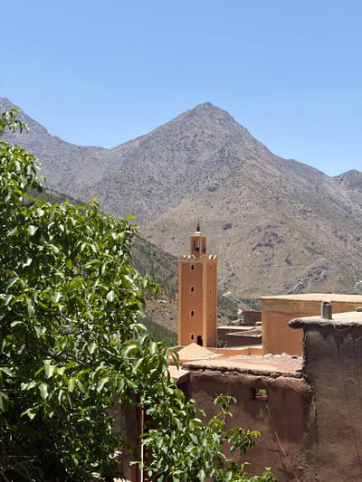 This image captures a mountainous landscape in Asni, Morocco, featuring traditional architecture under a clear blue sky. Dominating the background are arid, rugged mountains with sparse vegetation, extending across the entire horizon. A prominent peak rises in the upper central-right. The sky is uniformly clear and bright blue.

In the mid-ground, slightly left of center, a tall, square minaret stands out. It is constructed from an ochre-colored material, likely mud-brick or rammed earth, and features several small, rectangular window openings evenly spaced on its visible sides. The minaret's top is crenellated and surmounted by a finial. Further back on the mountain slopes to the right of the minaret, a winding dirt road is visible.

The foreground is occupied by dense, vibrant green foliage from a large tree, likely a walnut tree, which fills the left side of the frame and extends towards the center. Small green fruits are visible among the leaves. To the right and below this tree, several flat-roofed buildings made of reddish-brown, earthy materials are partially visible. A textured wall in the immediate bottom-right foreground has a small, square opening from which a thin wire extends downwards. No individuals or active subjects are present in the scene. The entire landscape is bathed in bright, direct sunlight.