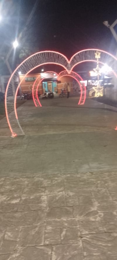 An outdoor night scene features illuminated heart-shaped arches creating a passageway. The arches have a wire-like structure with red lights outlining the hearts. In the background, there's a building, a decorative light display with star motifs, and parked motorcycles. The foreground showcases a stone-paved pathway leading towards the arches. Streetlights and faint figures are also visible, contributing to the ambiance.