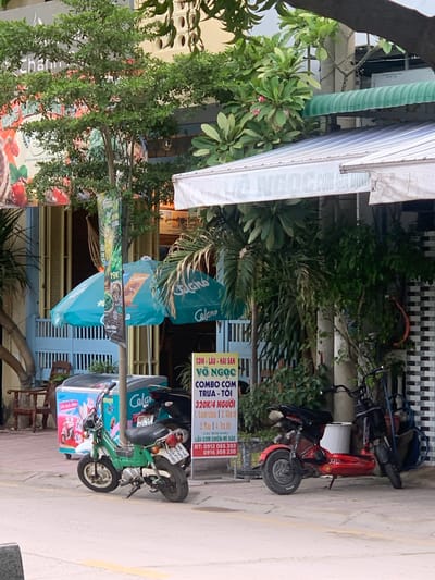 This is a medium shot, taken outdoors, of a street scene in Vĩnh Hải, Vietnam, with a focus on a few parked motorbikes and storefronts. The image is oriented vertically. In the foreground, a green and white motorbike is parked on a paved sidewalk next to a large, brightly colored cooler filled with what appear to be ice cream treats. A red motorbike is parked further back, behind a black-and-white checkered wall. Several trees and lush green plants are visible, with some in the foreground creating a dappled effect on the scene.
A large white awning extends from a building on the left, with text visible on its underside, partially obscured by a black tree branch. Below the awning, a sign with "Celano" logo and turquoise lettering is displayed. To the right of the motorbikes, a vertical sign provides menu information and contact details in Vietnamese. Text on this sign includes "COM-LAU HAI SAN", "VÕ NGỌC HI", "COMBO COM TRUA - TOI", and pricing information like "320K/4 NGƯỜI". Phone numbers are also listed: "0912 065 203" and "0916 308 230".
Another smaller sign, also with the "Celano" logo, is visible on the cooler. The overall atmosphere appears to be that of a casual street-side eatery or shop. The lighting suggests it could be daytime, possibly overcast or with the sun diffused by the awning and foliage. No people are clearly visible in the frame.