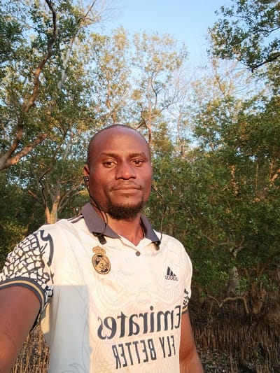 An outdoor image depicts a single adult male, positioned centrally, facing directly towards the camera, likely having taken a selfie due to the reversed text on his shirt. The individual has dark skin, a shaved head, and a short, trimmed beard. He is wearing a white short-sleeved polo shirt with a dark blue collar and trim, featuring a gold crest (Real Madrid logo) on the left chest and an Adidas logo on the right. The text "Emirates FLY BETTER" is visible in reverse on the front of the shirt. Black in-ear headphones are worn, with the cord visible around his neck. The subject maintains a neutral facial expression. The background consists of a dense, natural environment dominated by green foliage and numerous trees with visible, exposed roots at the lower right, indicative of a mangrove or swampy area. The sky above is clear blue, and the scene is illuminated by warm, natural light, suggesting either early morning or late afternoon. The location is consistent with a coastal or estuarine environment within Vanga ward, Kenya.