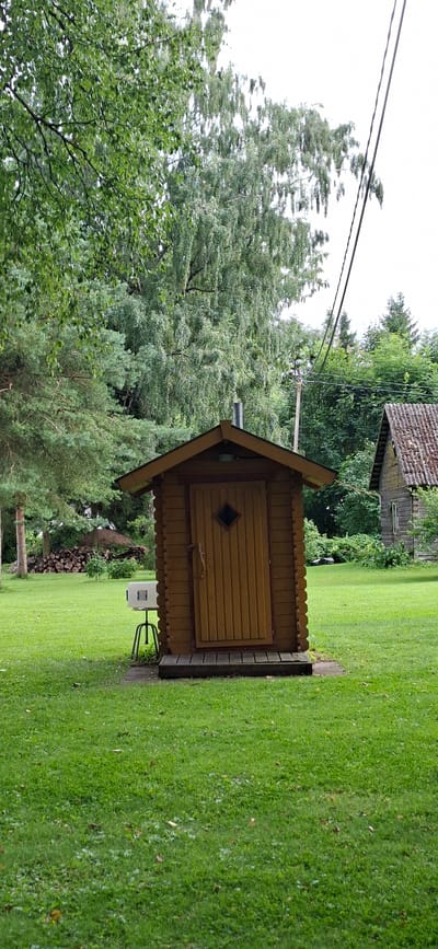 The image depicts an outdoor scene in Valma, Estonia, characterized by lush green grass and mature trees. Dominating the foreground is a small, rustic wooden shed with a pitched roof and a planked door featuring a diamond-shaped window. To the left of the shed, a white rectangular object with a metallic stand is visible. In the background, a pile of cut wood is stacked to the left, and a rustic wooden building with a tiled roof is situated to the right. Overhead, power lines stretch across the frame against a bright sky. The overall impression is one of a rural or semi-rural setting, possibly a backyard or a communal area.