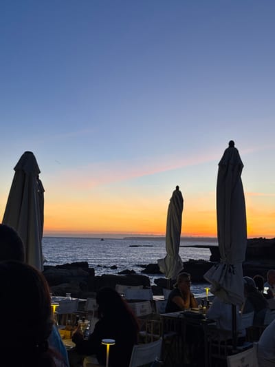 This image depicts an outdoor seaside dining establishment in Porches, Portugal, at dusk. The sky transitions from deep blue at the top to an orange-yellow sunset horizon, with faint pink hues. The ocean, dark blue with visible waves, extends from the rocky shoreline towards the distant horizon where a small boat is a dark silhouette.

In the foreground and mid-ground, multiple individuals are seated at tables in the open-air restaurant. Most people are in silhouette, but some details are discernible due to localized table lighting. One person with long dark hair is visible from behind in the lower left. To their right, another individual with long dark hair is seated, illuminated by a small, cylindrical table lamp. Further right, a person with lighter hair rests their chin on their hand, looking left. Other patrons are seated towards the right, mostly in silhouette.

The dining area features numerous tables and chairs, some with white fabric elements. Several items, including bottles, glasses, and illuminated table lamps, are visible on the tables. Large, folded light-colored patio umbrellas stand upright as prominent vertical silhouettes throughout the scene, with one on the far left, another near the center, and a third on the far right. The restaurant is situated directly on a dark, irregular rocky coastline that meets the sea.