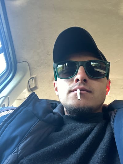 A man is seen from a low angle, wearing a black baseball cap, green sunglasses, and a dark-colored jacket, with a cigarette in his mouth. The setting appears to be the interior of a car, given the overhead interior fixtures and window visible in the background. The image does not provide any information regarding the location in the city of Biaga, Bulgaria.