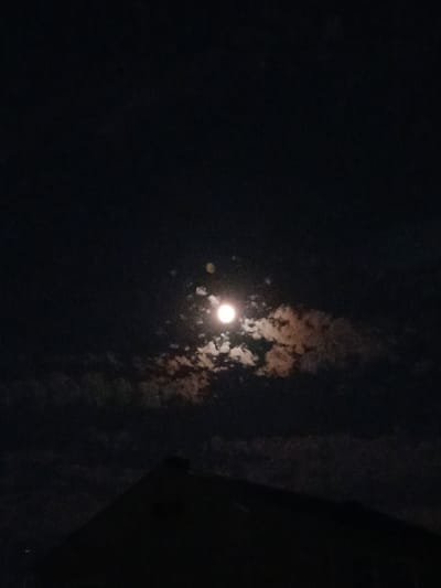 The image depicts a nighttime scene focused on the moon partially obscured by clouds. The moon is a bright, luminous orb in the center of the frame, casting a soft glow through the wispy, textured clouds that surround it. These clouds are a mix of white and brown tones, suggesting they are illuminated by the moonlight and possibly by some other light source, creating an ethereal effect.

The overall setting is dark, consistent with nighttime. The sky appears to be the primary subject, with the moon and clouds drawing the viewer's attention. To the right of the frame, a dark, angular shape is visible, possibly the edge of a building or a structure, contributing to the sense of an outdoor, terrestrial viewpoint.

There are no people or specific objects visible apart from the celestial bodies and the implied structure. The activity, if any, is passive – the observation of the moon and its interaction with the clouds.

Notable details include the time of day, clearly night. The weather is suggested by the presence of clouds, which are not dense enough to completely hide the moon, indicating a partly cloudy condition. The location context of Dinskaya, Russia, is provided, but there are no explicit visual cues within the image that directly point to this specific location, such as landmarks or distinctive features. The mood evoked by the image is one of quiet contemplation, perhaps a sense of mystery or natural beauty. No text is visible in the image.