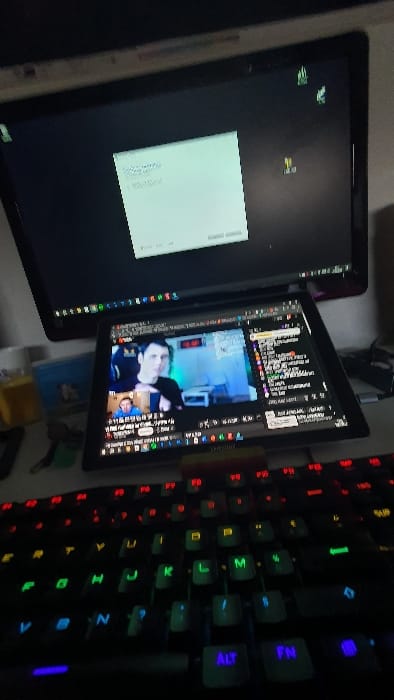 The image shows a desk in an indoor location, likely a home office or bedroom, with a computer monitor and a laptop. The monitor displays a message box, suggesting a computer issue or configuration process. The laptop screen shows a video stream of a man with a dark complexion and black hair, appearing to be a streamer or a participant in a video call.  The laptop and the keyboard are illuminated with a rainbow of colors, highlighting the keys, particularly the function keys and the "alt" key, which is partially visible. The keyboard's backlight is set to rainbow mode.  The location of the desk is in Arles, France, with coordinates 43.679 latitude and 4.598 longitude.  There is a small, yellow object on the desk, possibly a pencil or a charging cable. The desk is covered with a light-colored surface.  The image suggests a casual environment with an individual likely engaged in personal computing or streaming activities.