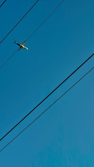 The media file captures an airplane flying in a clear blue sky, angled from a low perspective. Three thick, dark power lines diagonally traverse the frame from the bottom left to the top right. The airplane, white with yellow and blue accents, is positioned in the lower left quadrant, between the two uppermost power lines. Its landing gear is down, suggesting it is either taking off or landing. The bright, clear sky indicates it is daytime, likely sunny. No people are visible. The scene is set against a vast expanse of sky, with the power lines providing a strong geometric element. The presence of power lines suggests an area with infrastructure, potentially near an airport or a town. The overall mood is one of openness and travel, with the airplane serving as the focal point against the backdrop of the sky and man-made structures. There is no visible text on the airplane or in the surrounding environment. The location is Piatra Albă, Moldova.