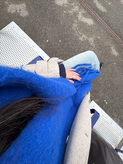 Overhead shot of a person seated outdoors. The person wears blue jeans, a navy sweater, and a light gray coat. A bright blue, fluffy scarf is draped across their lap. The person's left hand, adorned with multiple rings, rests on the scarf. The person sits on a silver, perforated bench with a blue accent. A dark bag sits to the upper right. A concrete surface is visible to the left, with a linear drainage grate. Some strands of long, dark hair are visible beneath the scarf.