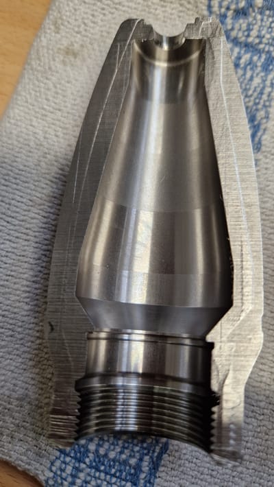 This is a close-up, cutaway photograph of a metal object, likely a component of machinery or a firearm accessory, resting on a textured, blue and white striped cloth. The object is metallic and appears to be made of machined steel, exhibiting a brushed or satin finish with visible tool marks and concentric rings from the manufacturing process.

The object is cylindrical and appears to be a hollow tube with a tapering interior. One end of the object is open and features intricate threading, suggesting it is designed to screw onto another component. The other end is narrower and has a more complex, stepped internal shape, with a rounded inner edge. There are some light scratches and imperfections visible on the metal surface, particularly on the outer casing.

The lighting suggests an indoor setting, with a soft, diffused light source casting subtle reflections on the polished metal. No people are visible in the image. The background is composed of the cloth and a hint of a wooden surface below it. The time of day and weather are not discernible. There is no visible text in the image. The overall impression is one of a precisely engineered mechanical part, viewed in a functional or informational context, possibly for inspection or demonstration. The location context of Ichenhausen, Germany, does not provide specific visual cues within the image itself.