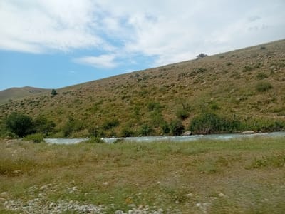 The image depicts a landscape scene with a sloping, dry, grassy hillside dotted with rocks and sparse green vegetation. A body of flowing water, likely a river or stream, runs horizontally across the lower portion of the frame. The water appears to be a light blue color and is in motion, with some ripples visible. The foreground consists of dry, patchy grass and pebbles. In the background, the hillside continues upwards towards a hazy sky with scattered white clouds. The overall impression is one of a natural, undeveloped environment, possibly in a rural or mountainous region. The location is specified as Көксаяқ ауылдық округі, Kazakhstan. There are no people or animals visible in the image, nor any man-made structures or activities.