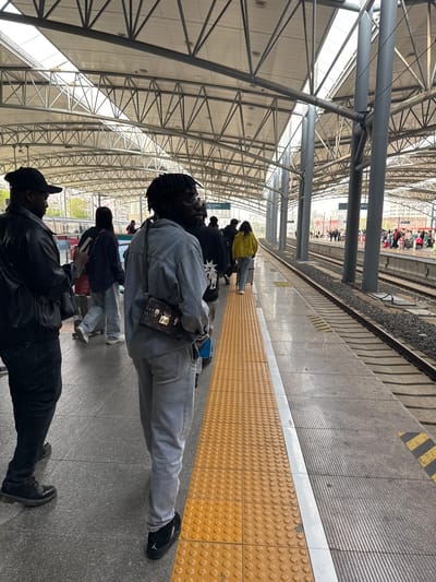 The image depicts a railway platform, presumably at a train station, in Shenhe District, China. Several people are standing or walking on the platform, alongside a train track. The platform is marked with yellow tactile paving, indicating the edge. The station has a large, arched, metal-framed roof structure. People are wearing casual clothing. One individual is in a light denim jacket and jeans, with a crossbody bag. Another is wearing a black leather jacket. The overall scene suggests a typical train station environment with people waiting or passing through.
