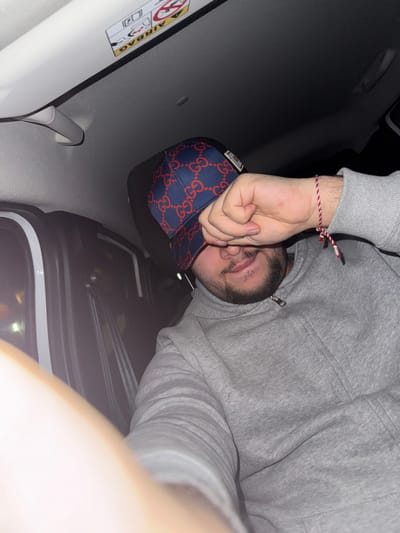 The image depicts a male individual seated in the passenger side of a vehicle, appearing to be at an unspecified location within Vellmar, Germany. The male subject is observed from a slightly low angle, positioned towards the right side of the frame, partially obscuring his face with his left hand. He has a dark beard and mustache, and his mouth is subtly curved in what appears to be a slight smile. He is wearing a grey zippered hooded sweatshirt and a dark blue baseball cap with a red "GG" (Gucci) pattern, worn backward. On his left wrist, a red and white braided string bracelet is visible.
The immediate environment is the interior of a car. The white ceiling panel of the vehicle is visible in the upper half of the image, featuring a grey grab handle on the passenger side. An "AIRBAG" warning sticker, displaying German text and international symbols indicating airbag and child seat hazards, is affixed to the sun visor. The vehicle's dark-colored interior upholstery is partially visible behind the subject. To the left side of the frame, part of a white car door frame and a dark window are visible, beyond which blurry, yellow-white external light sources suggest an outdoor, low-light, possibly nighttime setting. The subject's right arm and torso occupy the lower portion of the image.