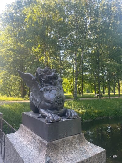 The image depicts a detailed, eye-level view of a winged mythical creature statue set against a lush park backdrop, located in Mar'ino, Russia. In the foreground, a substantial, dark-patinated cast-iron statue, resembling a griffin or winged lion, rests on a dark rectangular plinth. The statue features prominent, feathered wings, an open mouth with an extended tongue, clawed forepaws, and a circular, ornate emblem on its side. This plinth is situated atop a large, light-grey, mottled stone pedestal, likely granite, with a decorative curved edge at its top. To the immediate left of the pedestal, a dark metal railing, possibly part of a bridge or embankment, is partially visible.

To the right of the pedestal, a calm body of dark water, likely a canal or pond, occupies the lower right quadrant of the image. Subtle ripples and reflections of the sky and foliage are visible on its surface. A single mallard duck, identifiable by its green head and mottled brown body, swims towards the right edge of the frame.

The mid-ground and background are dominated by a verdant park scene. A wide, light-colored pathway, appearing unpaved, stretches horizontally across the mid-background. Beyond the path, an abundance of mature deciduous trees with vibrant green foliage forms a dense canopy. Several birch trees with distinctive white bark are scattered among the other trees. Sunlight filters through the leaves, creating bright highlights and dappled light effects throughout the park. On the pathway, in the background to the right of the statue, two individuals are visible; one is seated on a light-colored bench, and another stands nearby. Their features are indistinct due to distance. The overall scene is brightly lit by natural sunlight, suggesting a clear day during spring or summer.