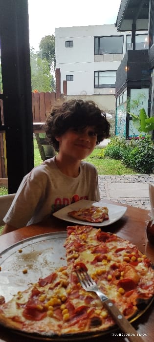 The scene depicts a young male at an outdoor dining establishment in Tumbaco, Ecuador, at approximately 16:25 on February 27, 2026. A light-skinned male with dark, curly hair, wearing a light-colored t-shirt, is seated at a wooden table, smiling and looking towards the viewer. In the foreground, a large, partially consumed pizza rests on a round metal pan. The pizza features toppings including processed meat (appears to be pepperoni or ham) and corn kernels. A metal fork with a wooden handle is embedded in a slice of pizza on the pan. To the male's right, a single slice of pizza is presented on a white rectangular plate. The background reveals an outdoor setting with a landscaped garden area, a paved path, and several buildings. A two-story, light-colored building with dark-framed windows is visible behind the garden, and a darker-colored building with large glass sections is visible to the right. A wooden fence and trees are also present in the background.