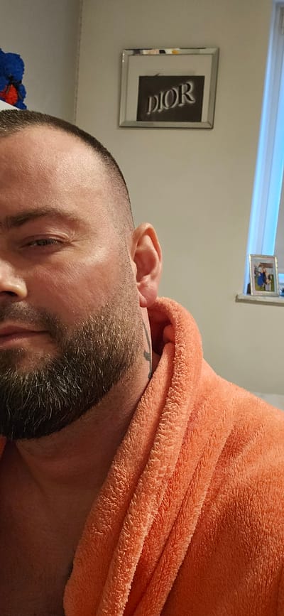 A man is wearing a coral towel around his neck. He has a beard and a tattoo on his neck. Behind him is a wall with a framed DIOR print and another framed photo. The location is implied to be Gütersloh, Germany.