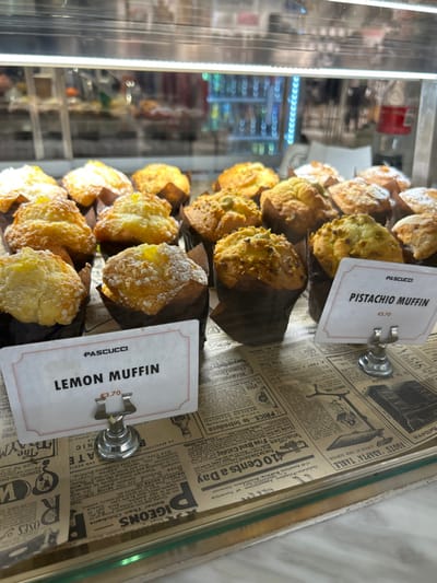 The image depicts a close-up view inside a glass display case at a Pascucci café, located in Saint Julian's, Malta. Approximately thirty baked muffins are arranged across two visible rows. The foreground features several "Lemon Muffin" varieties, distinguished by a light yellow glaze and a dusting of powdered sugar, displayed in dark brown paper liners. One white price tag, labeled "PASCUCCI LEMON MUFFIN €3.70," is positioned on the left. To the right, "Pistachio Muffin" varieties are visible, characterized by a light brown exterior topped with chopped nuts, also in dark brown paper liners. A second white price tag, labeled "PASCUCCI PISTACHIO MUFFIN €3.70," is positioned on the right. Both price tags are held by small, metallic stands. The base of the display case is covered with paper resembling vintage newspaper print. The background, out of focus, shows reflections of a brightly lit interior, including what appears to be a refrigerated display with blue lighting and various beverages or products. No individuals are visible.